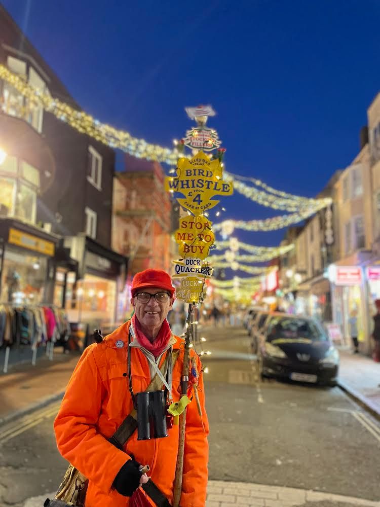 The birdyman, a white man wearing glasses, an orange jacket and hat, binoculars and holding a pole adorned with signs advertising his bird whistles, smiles at the camera. 