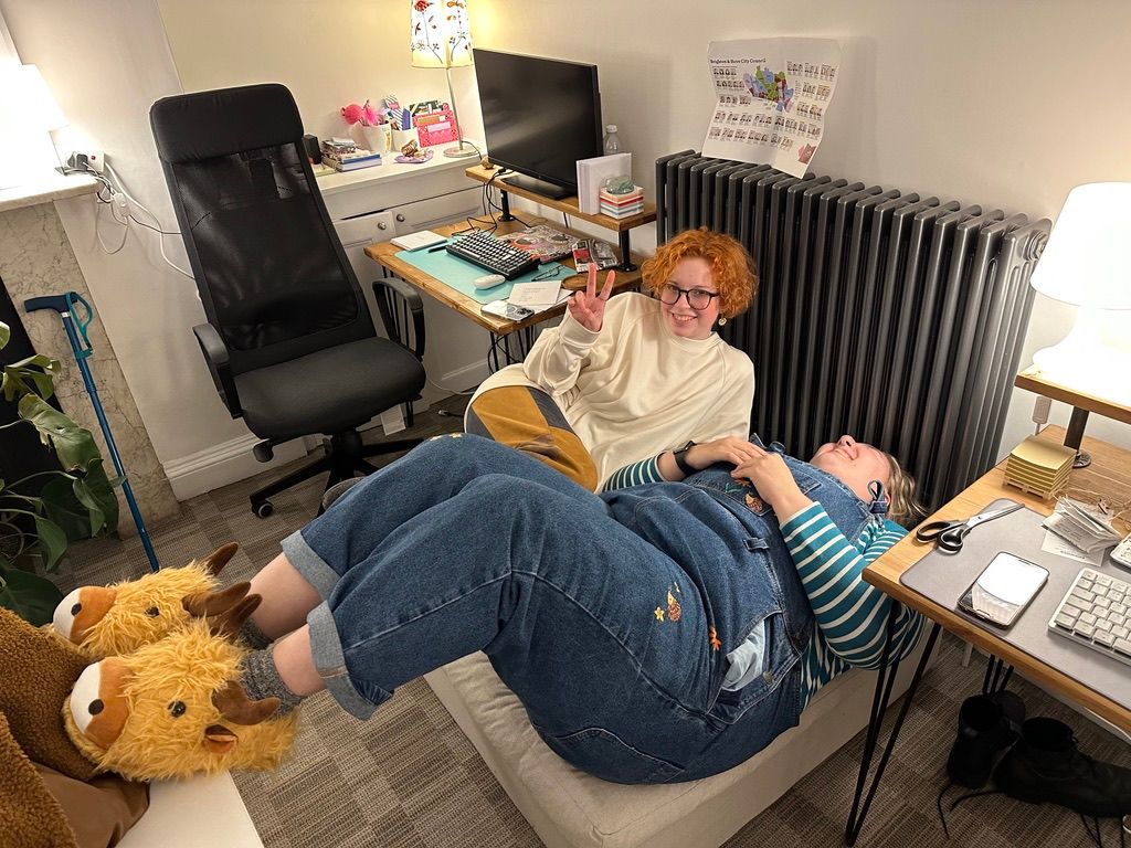 Two members of Seagull in an office, sat on a square footstool. One person with ginger hair, a cream top and black glasses is throwing a peace sign. The other is lying down, wearing dungarees with hedgehogs on and a blue and white stripy top, with highland cow slippers. They are both smiling.