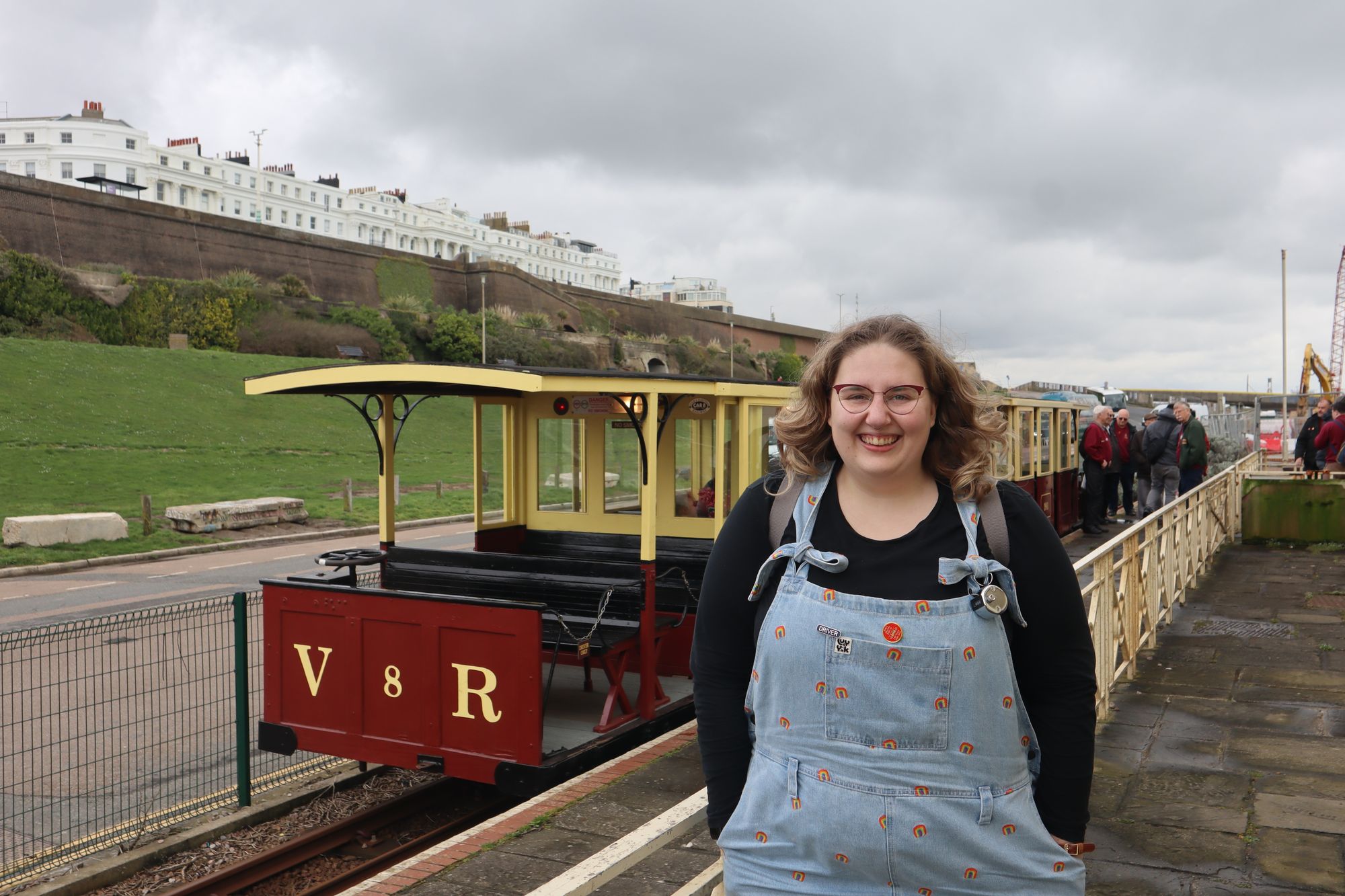 A photo of Carly-May, wearing denim dungarees with embroidered rainbows on, in front of car '8' of the Volk's Electric Railway at Black Rock Station.