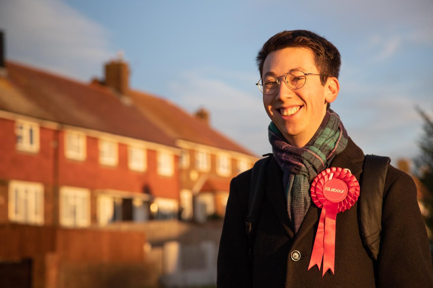 A photo of Jacob Allen, a white man with round glasses and short brown hair, wearing a black coat, tartan scarf and Labour rosette, smiling in front of houses.