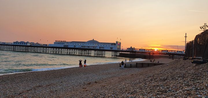 Palace Pier at sunset, from the beach in Kemptown. The sky is orange and you can see the i360 in the distance. 