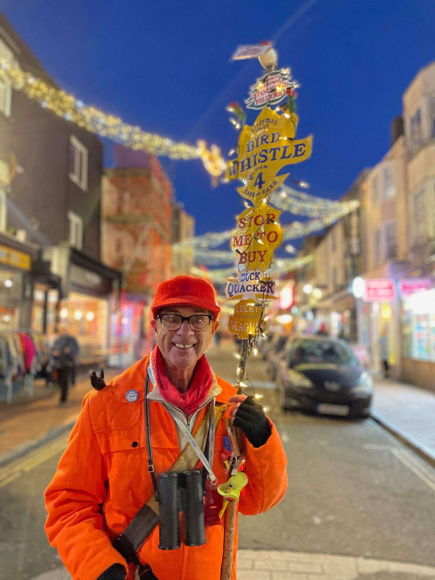 The birdyman, a white man wearing glasses, an orange jacket, binoculars and holding a pole adorned with signs advertising h