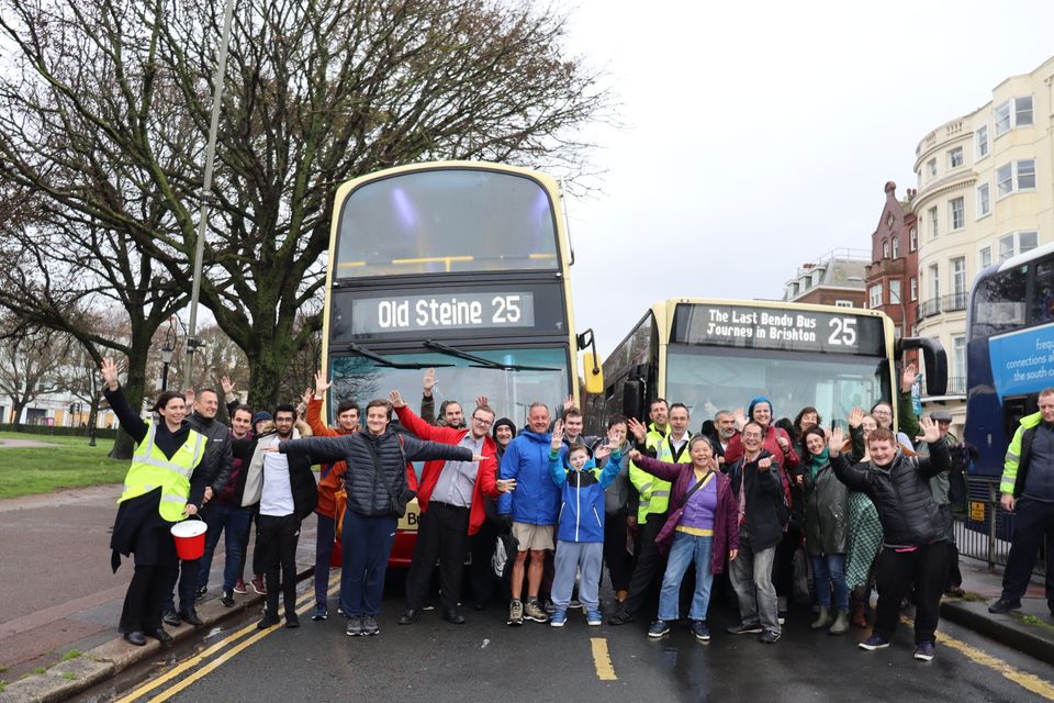Two dozen bus fans posing in front of a double decker 25 and a bendy bus.